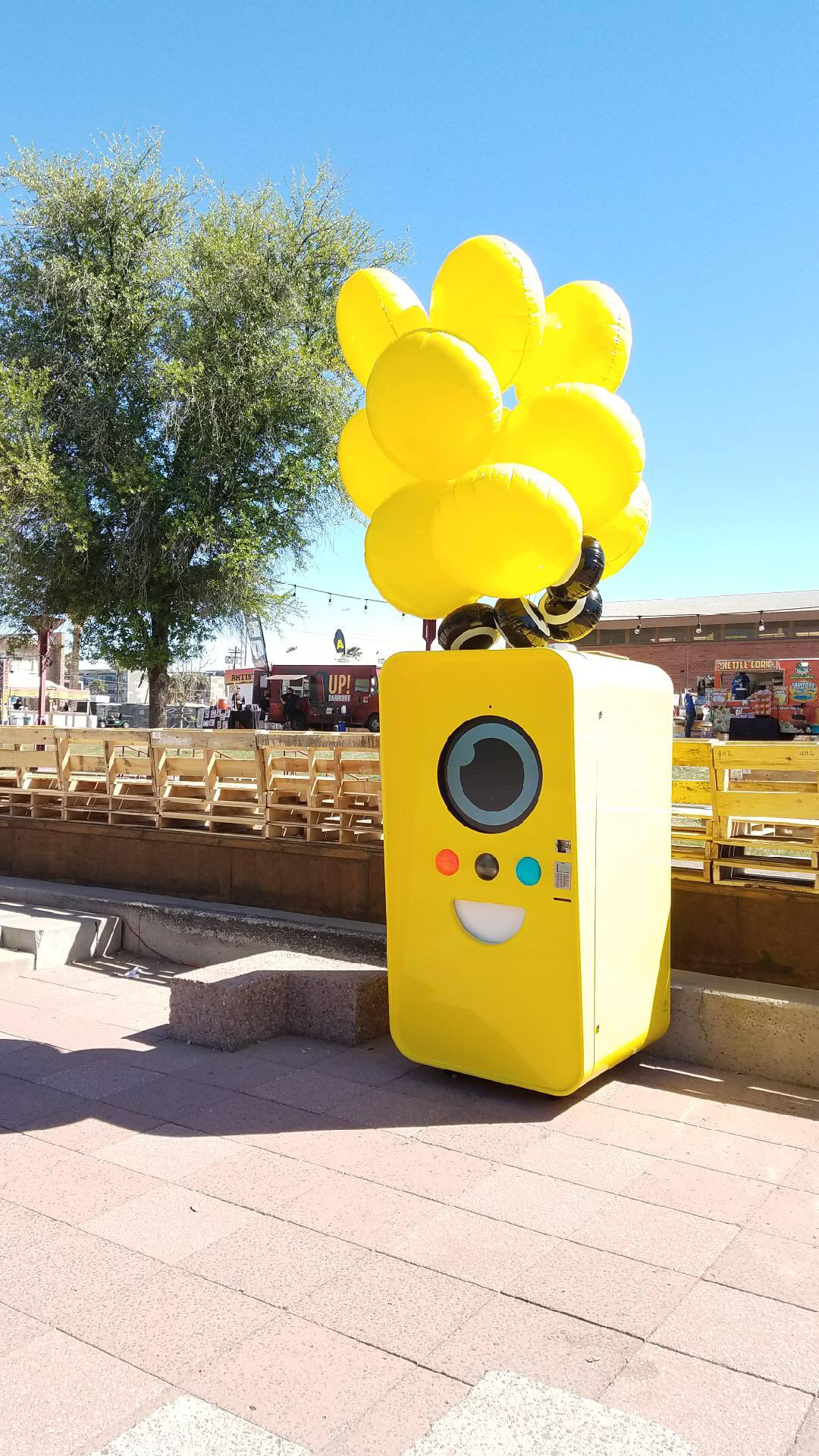 Yellow Snapbot vending machine with balloons on a paved plaza beside wooden bleachers, food trucks and a kettle corn stand